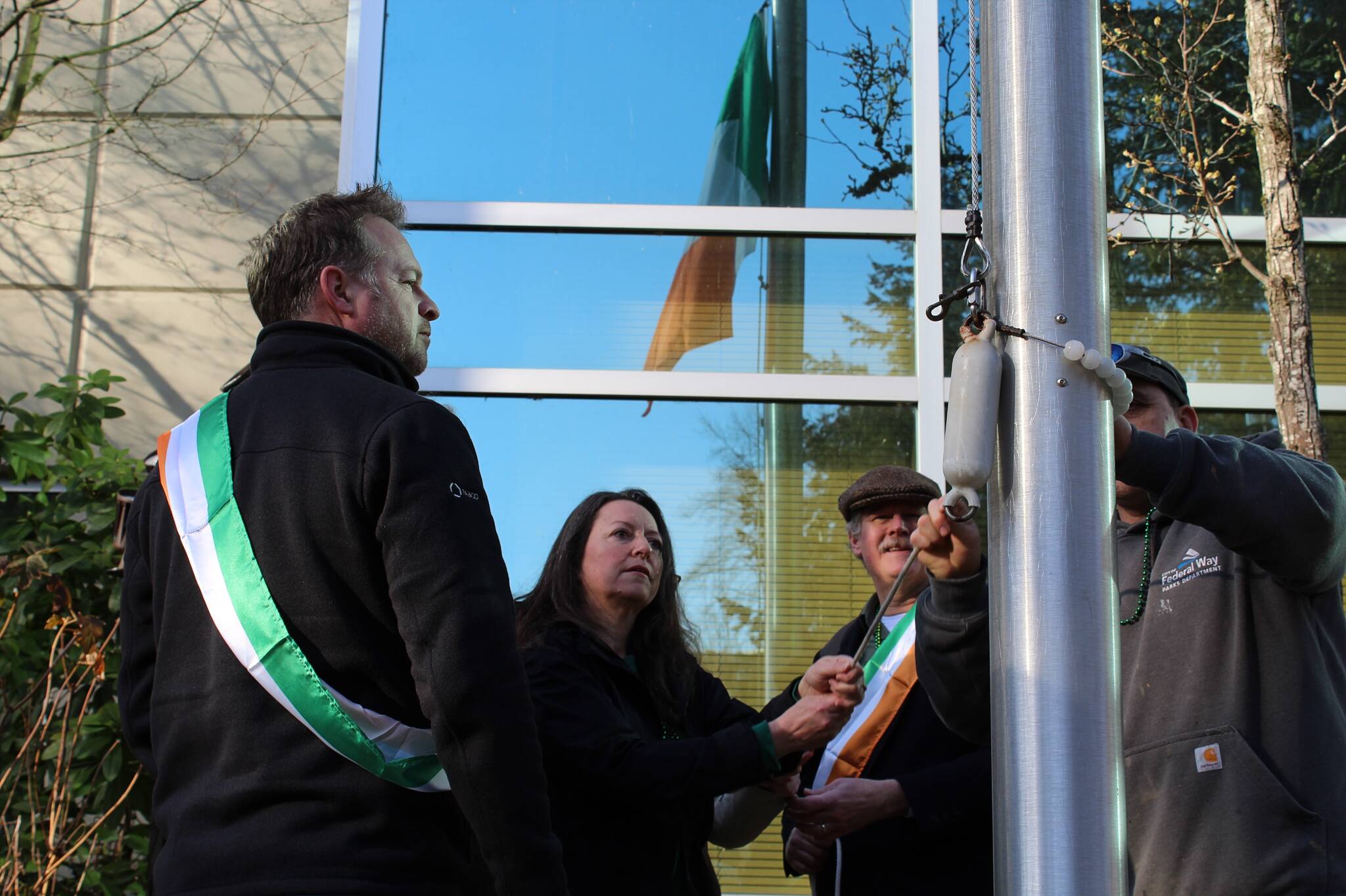 The Patrick family, friends and a city employee raise the Irish flag, as visible in the reflection of City Hall. Alex Bruell / The Mirror