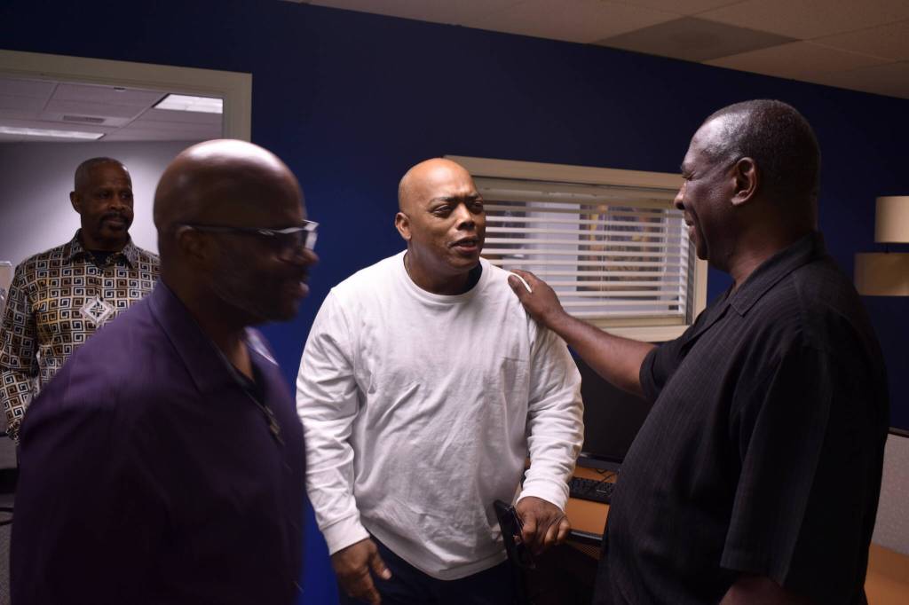 Pastor George Merriweather places his hand on the shoulder of Pastor Charles Allen, who is an Empowered Youth Project Mentor, during a conversation. Curtis Retic, board chair of the Good Shepherd Youth Outreach, stands in the doorway, and Louis Guiden walks by the group.
