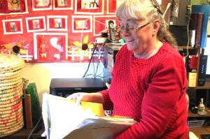 Local author Teresa Bateman holds a thick folder of her personal antidotes for writers block. The folder is full of her ideas and inspiration. Behind her is a large red poster that has hung on the wall behind her desk for years. It was made by some of her students and based on one of her picture books. Photo by Keelin Everly-Lang/ The Mirror.
