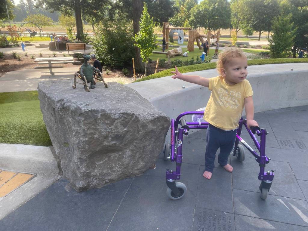 A child with a walker points to a fun statue of an animal using a walker at the Chelsea Anderson Memorial Play Station (CHAMPS) @Marshall Park in Vancouver, Washington. Photo from Harpers Park, the organization that designed, funded and provided construction oversight for the playground. Concept design for the park was by Todd Girvin of Harpers Playground.