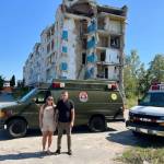 Vitaliy and Irina Piekhotin in Ukraine in front of the two ambulances they delivered and a building that has been bombed. Photo provided by Vitaliy Piekhotin.