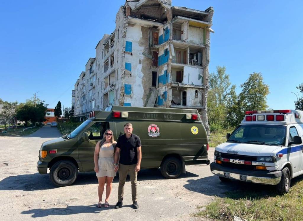 Vitaliy and Irina Piekhotin in Ukraine in front of the two ambulances they delivered and a building that has been bombed. Photo provided by Vitaliy Piekhotin.